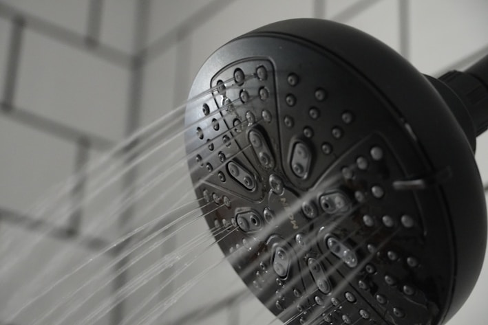 A black showerhead with multiple tiny openings is mounted against a tiled wall. Water streams are visibly spraying out in several directions, capturing a dynamic aspect of flowing water.