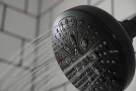 A black showerhead with multiple tiny openings is mounted against a tiled wall. Water streams are visibly spraying out in several directions, capturing a dynamic aspect of flowing water.