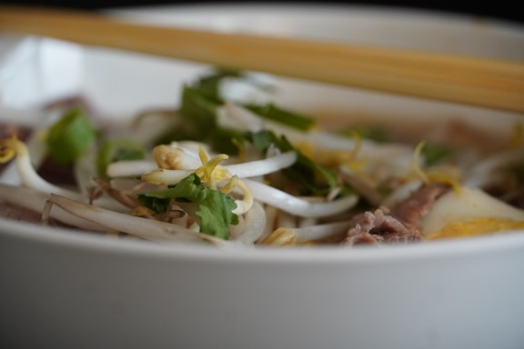 A close-up view of a bowl of Vietnamese pho, featuring fresh bean sprouts, slices of beef, green herbs, and pieces of lime. The dish is arranged in a white bowl, with wooden chopsticks resting on the rim.