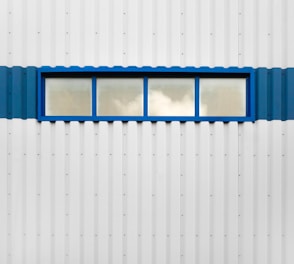 A rectangular blue-framed window is set in the middle of a corrugated metal wall. The window is divided into four panes, with a reflection of clouds visible in the glass. The wall itself is predominantly white with a blue stripe running horizontally along the middle where the window is situated.