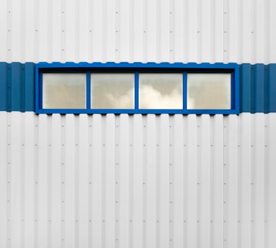 A rectangular blue-framed window is set in the middle of a corrugated metal wall. The window is divided into four panes, with a reflection of clouds visible in the glass. The wall itself is predominantly white with a blue stripe running horizontally along the middle where the window is situated.
