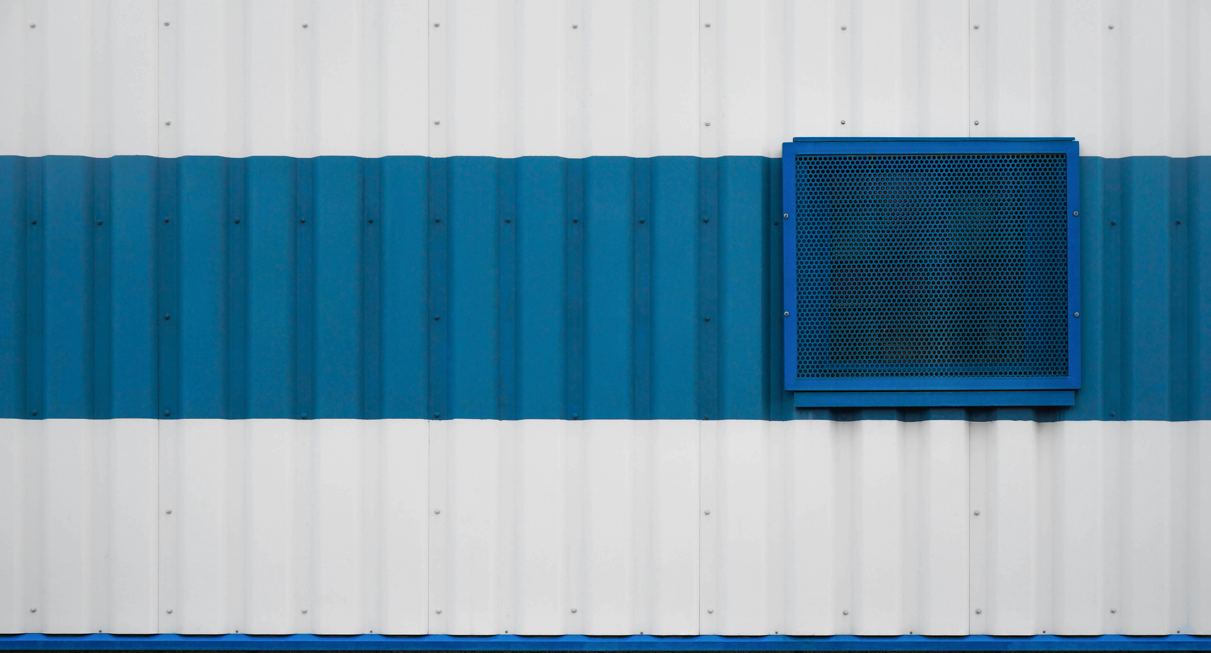 A corrugated metal wall with alternating blue and white horizontal stripes. A blue square vent with a mesh detail is prominently featured on the right side of the wall.