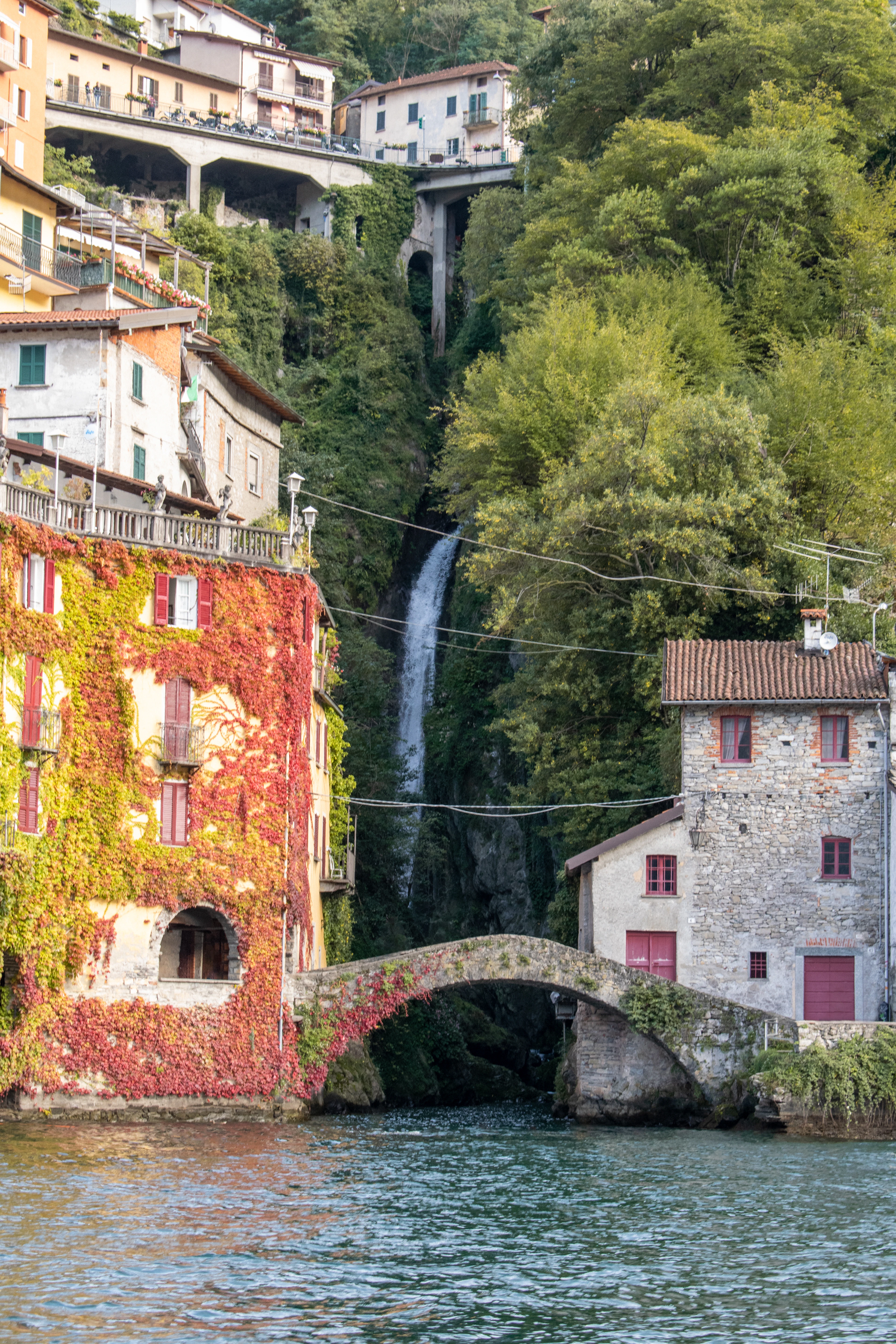 The old stone arch bridge at Nesso on Lake Como spanning the mouth of the Orrido gorge, a slender waterfall dropping through the trees behind it and an autumn-red creeper covering the tall house on the left, above the green lake water.