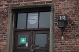 A brick building facade with a window and a door. The door has a sign that reads 'THE CITIZEN BETRAYED TO THE MEMORY OF THE HUNGARIAN HOLOCAUST' in English and another language. A lampshade with the number 18 is above the door. A green and white medical cross symbol is on one of the glass panes of the door.