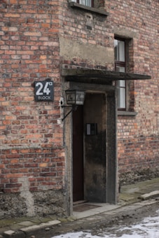 An old brick building wall with a small door and a sign reading '24a Block' beside a lantern. The bricks are weathered and the ground has a mix of snow patches and stone paving.