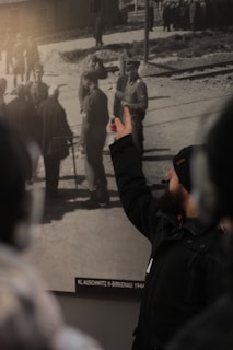 A person wearing a headset and winter clothing gestures toward a historic black-and-white photograph displayed on a wall. The photograph depicts a group of people, including military personnel and civilians, in an outdoor setting with a railway track visible.