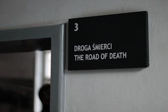 A black sign mounted on a white wall displaying the number three and the text 'DROGA ŚMIERCI THE ROAD OF DEATH' in white letters.