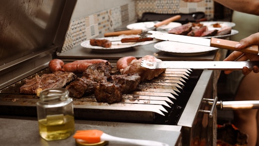A variety of meats are grilling on an outdoor barbecue. There are sausages and large cuts of steak on the grill, with tongs being used to handle the meat. A jar of oil and a brush are on the counter beside the grill, and plates with additional pieces of meat sit in the background.