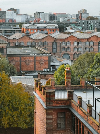 Multiple brick buildings with industrial architecture are surrounded by greenery, set against a backdrop of a cityscape with modern buildings in the distance. The words 'Manchester Studios' are visible on one of the buildings.
