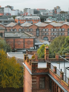Multiple brick buildings with industrial architecture are surrounded by greenery, set against a backdrop of a cityscape with modern buildings in the distance. The words 'Manchester Studios' are visible on one of the buildings.
