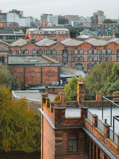 Multiple brick buildings with industrial architecture are surrounded by greenery, set against a backdrop of a cityscape with modern buildings in the distance. The words 'Manchester Studios' are visible on one of the buildings.