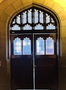 A pair of ornate wooden doors adorned with intricate stained-glass windows, set within an arched stone doorway. The stained glass features geometric and floral patterns, with a mix of colors including blue, purple, and white. The stone framework surrounding the door has a smooth finish and a warm, yellowish tone.