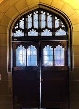 A pair of ornate wooden doors adorned with intricate stained-glass windows, set within an arched stone doorway. The stained glass features geometric and floral patterns, with a mix of colors including blue, purple, and white. The stone framework surrounding the door has a smooth finish and a warm, yellowish tone.