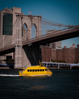 A bright yellow water taxi glides across a river in front of the iconic Brooklyn Bridge. The bridge's stone arches and suspension cables are prominently visible, with city skyscrapers and a building with clear signage in the background. The American flag waves atop a structure, all under a clear blue sky.