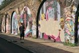 A person stands in front of a series of graffiti-covered walls, possibly under an overpass or archway. The walls are adorned with colorful and varied street art, including stylized portraits and abstract designs. The person appears to be observing or contributing to the artwork. Sunlight casts shadows, enhancing the vibrant colors of the art.