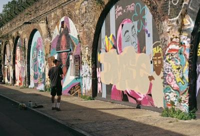 A person stands in front of a series of graffiti-covered walls, possibly under an overpass or archway. The walls are adorned with colorful and varied street art, including stylized portraits and abstract designs. The person appears to be observing or contributing to the artwork. Sunlight casts shadows, enhancing the vibrant colors of the art.