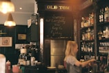 A cozy bar interior with warm lighting, featuring a wooden counter filled with bottles and a bartender preparing drinks. The walls are decorated with framed artwork and a small chalkboard displaying writing. An old-fashioned vibe is conveyed by signs and decor.