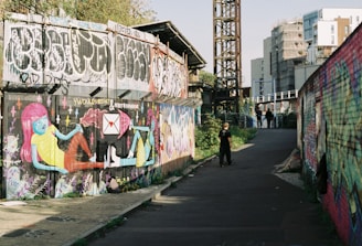 A vibrant street art mural covers a section of a wall, depicting colorful characters against a backdrop of graffiti. The scene is set in an urban alleyway, with both colorful and monochrome graffiti on the nearby surfaces. A metal structure and modern buildings can be seen in the distance. People are walking along the path, adding a sense of movement and life to the otherwise static artwork.