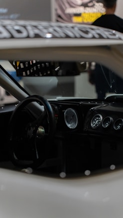 Interior of a car featuring a steering wheel, a dashboard with multiple gauges, and a racing seat. The focus is on the dashboard, which includes analog dials, suggesting a classic or racing car design. The lighting is dim, contributing to a sleek and serious ambiance.