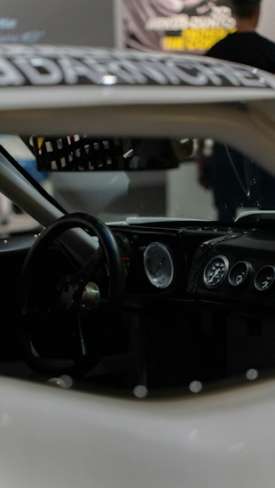 Interior of a car featuring a steering wheel, a dashboard with multiple gauges, and a racing seat. The focus is on the dashboard, which includes analog dials, suggesting a classic or racing car design. The lighting is dim, contributing to a sleek and serious ambiance.