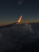 An airplane wing is visible against a backdrop of a dark sky transitioning to sunset hues. Clouds are significantly visible below the wing, illuminated subtly by the setting sun. The tip of the wing displays branding, with the logo reflecting the sunlight.