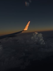 An airplane wing is visible against a backdrop of a dark sky transitioning to sunset hues. Clouds are significantly visible below the wing, illuminated subtly by the setting sun. The tip of the wing displays branding, with the logo reflecting the sunlight.