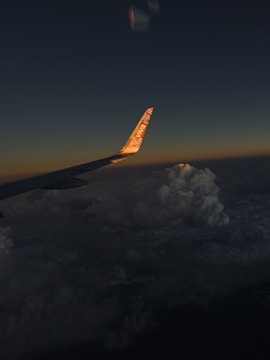 An airplane wing is visible against a backdrop of a dark sky transitioning to sunset hues. Clouds are significantly visible below the wing, illuminated subtly by the setting sun. The tip of the wing displays branding, with the logo reflecting the sunlight.