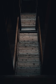 A wooden staircase viewed from above, surrounded by dim lighting and shadows. The steps are worn and display a natural wooden texture, framed by wooden railings on either side.
