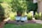 A lush garden scene with a small tree growing from a cut stump surrounded by vibrant white and purple flowers in pots. The garden is well-maintained, featuring green grass and paved stone paths. Hedges and a few larger shrubs create a natural border, while a white chair and other garden furniture are partially visible in the background.