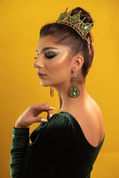 A woman with wavy brown hair is adorned with a tiara featuring green gemstones. She wears elaborate, matching green earrings and a green velvet dress. Her makeup is bold, with dramatic eye shadow and contouring. The background is a solid, vibrant yellow.