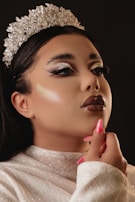 A close-up of a person wearing a sparkling silver crown. They have a glamorous makeup look with shimmering eyeshadow, thick eyelashes, glossy lipstick, and their hair is styled elegantly. The person is dressed in a glittery white top, touching their chin with manicured pink nails. The background is dark, highlighting the facial features and crown.