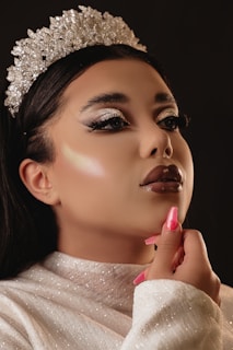 A close-up of a person wearing a sparkling silver crown. They have a glamorous makeup look with shimmering eyeshadow, thick eyelashes, glossy lipstick, and their hair is styled elegantly. The person is dressed in a glittery white top, touching their chin with manicured pink nails. The background is dark, highlighting the facial features and crown.