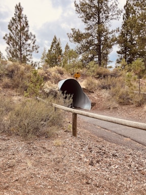A large corrugated metal pipe embedded into the ground serves as a tunnel under a dirt path. Tall pine trees and brush surround the area, giving it a natural, forest-like environment. A wooden rail runs along the path, and a yellow caution sign is visible above the tunnel entrance.