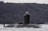 A military submarine partially submerged in water, with a few sailors standing on top. The vessel is navigating through a large body of water against a backdrop of a wooded hillside. A flag is visible on the submarine.