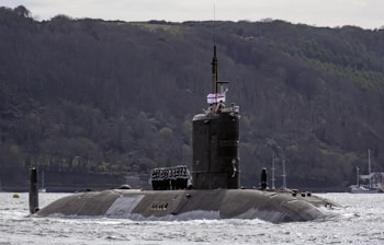 A military submarine partially submerged in water, with a few sailors standing on top. The vessel is navigating through a large body of water against a backdrop of a wooded hillside. A flag is visible on the submarine.