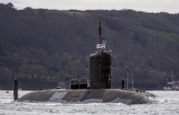 A military submarine partially submerged in water, with a few sailors standing on top. The vessel is navigating through a large body of water against a backdrop of a wooded hillside. A flag is visible on the submarine.