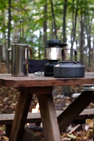 A rustic wooden picnic table is set up in a forested area with trees in the background. On the table, there is a metal French press, a black camping lantern, and a metal kettle. The ground is covered with fallen leaves, and a small campfire can be seen underneath the table.