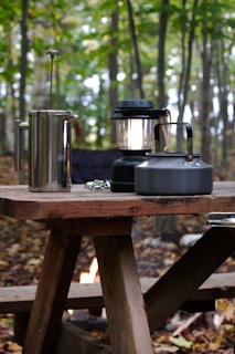 A rustic wooden picnic table is set up in a forested area with trees in the background. On the table, there is a metal French press, a black camping lantern, and a metal kettle. The ground is covered with fallen leaves, and a small campfire can be seen underneath the table.