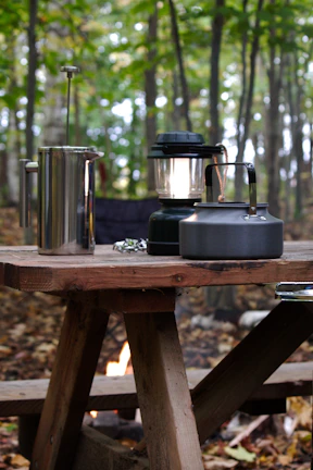 A rustic wooden picnic table set with colorful Campora camping plates and mugs under a canopy of pine trees.