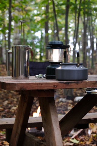 A rustic wooden picnic table is set up in a forested area with trees in the background. On the table, there is a metal French press, a black camping lantern, and a metal kettle. The ground is covered with fallen leaves, and a small campfire can be seen underneath the table.