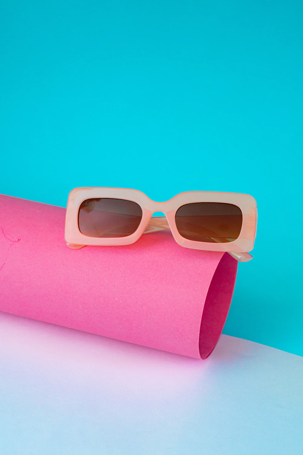 Milky blush rectangular sunglasses balanced on a rolled pink sheet against an aqua wall