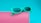 A pair of vibrant turquoise sunglasses rests on a pink, cylindrical surface. The background is a solid turquoise color, creating a bold contrast with the pink. The sunglasses have a modern, slightly oversized frame with dark lenses.
