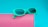 A pair of vibrant turquoise sunglasses rests on a pink, cylindrical surface. The background is a solid turquoise color, creating a bold contrast with the pink. The sunglasses have a modern, slightly oversized frame with dark lenses.