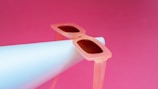 A pair of stylish, translucent pink sunglasses rests atop a white cylindrical object, set against a vibrant pink background. The sunglasses have a retro feel, with oversized frames and dark lenses.
