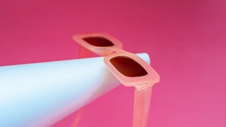 A pair of stylish, translucent pink sunglasses rests atop a white cylindrical object, set against a vibrant pink background. The sunglasses have a retro feel, with oversized frames and dark lenses.
