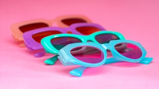 Several pairs of colorful sunglasses are laid out against a bright pink background. The frames of the sunglasses are in playful pastel shades, including purple, turquoise, and peach, giving off a retro and summer vibe.