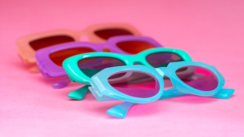 Several pairs of colorful sunglasses are laid out against a bright pink background. The frames of the sunglasses are in playful pastel shades, including purple, turquoise, and peach, giving off a retro and summer vibe.