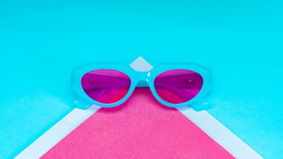 A pair of sunglasses with a light blue frame and pink-tinted lenses lies on a geometric background of vibrant blue and pink shades. The eyewear is centered and stands out against the contrasting colors, creating a modern and stylish aesthetic.