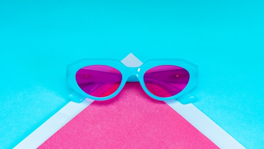 A pair of sunglasses with a light blue frame and pink-tinted lenses lies on a geometric background of vibrant blue and pink shades. The eyewear is centered and stands out against the contrasting colors, creating a modern and stylish aesthetic.
