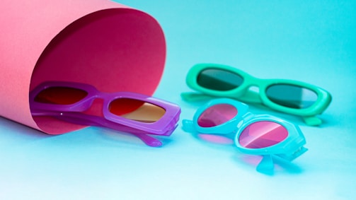 A collection of colorful sunglasses is arranged on a blue surface. The sunglasses are in vibrant hues, including purple, turquoise, and teal. A large pink paper cylinder partially covers one pair, adding a playful and bright contrast to the scene.